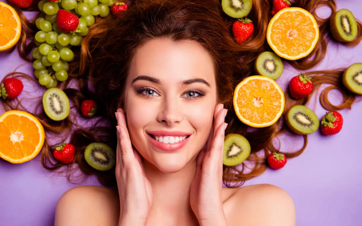 A woman with long brown hair smiles while surrounded by various fruits including orange halves, kiwi slices, strawberries, and green grapes, all arranged on a purple background. She is touching her cheeks with both hands. Superfoods for Glowing Skin