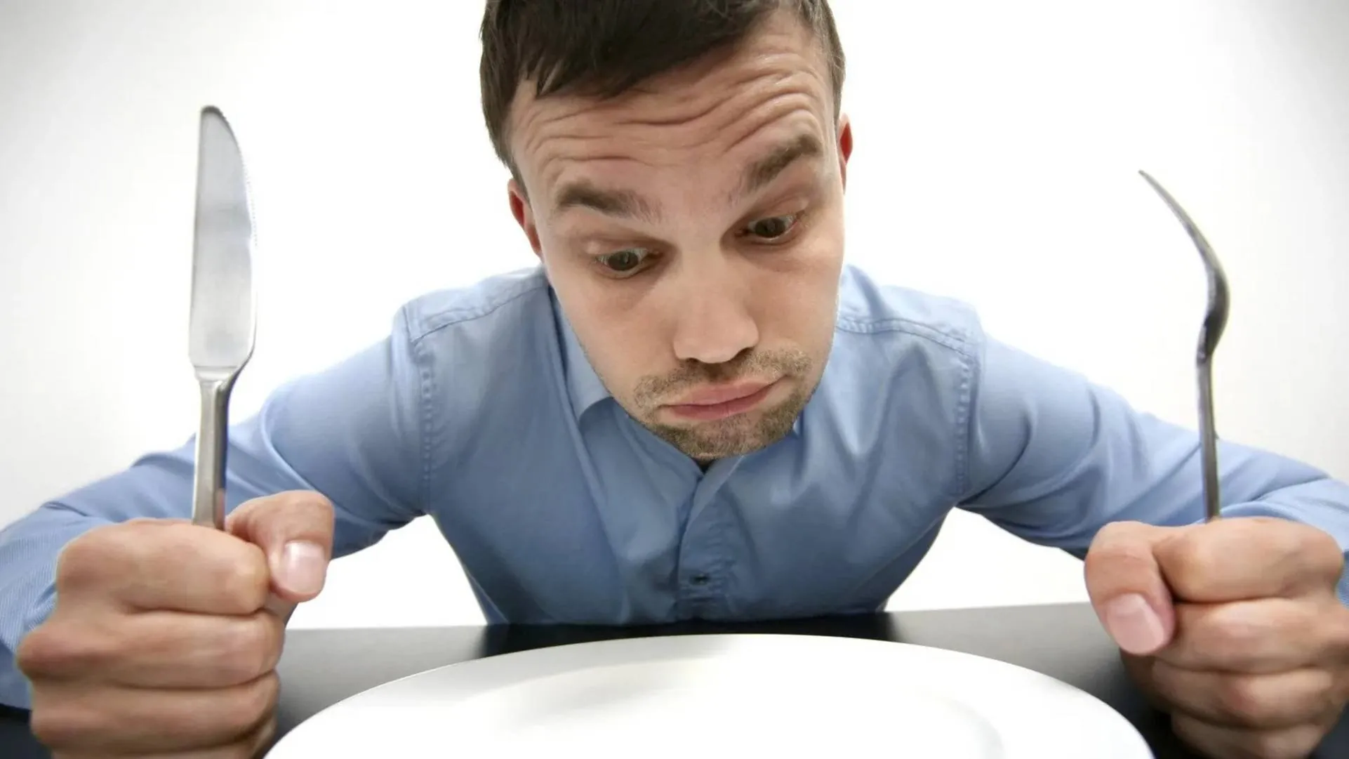 A man in a blue shirt sits at a table, holding a knife and fork, looking disappointedly at an empty white plate.