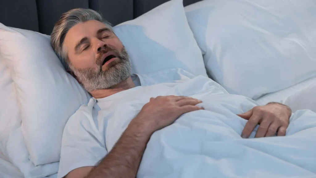 A middle-aged man with gray hair and a beard is lying in bed, sleeping on his back with his mouth open and one hand resting on his chest, covered by a white blanket.