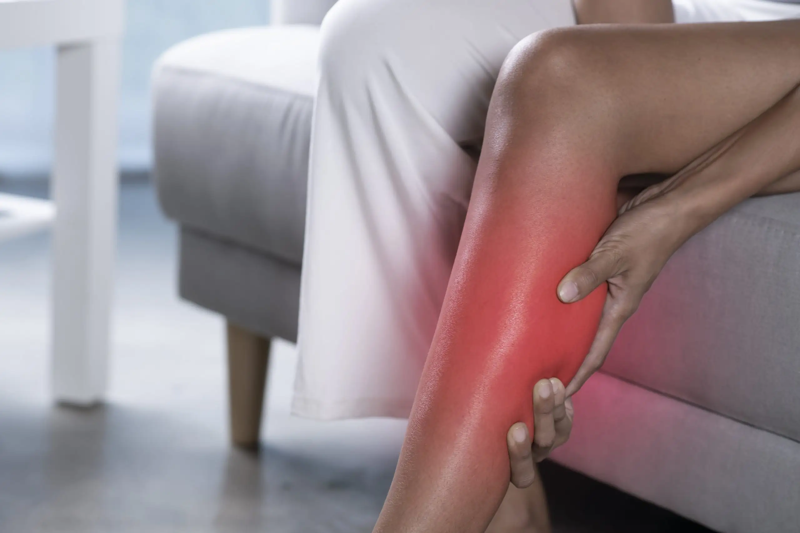 What Kind of Magnesium for Leg Cramps Works Fast 3 A person sitting on a couch holds their lower leg, which is highlighted in red to indicate pain or discomfort, possibly from a cramp or injury.